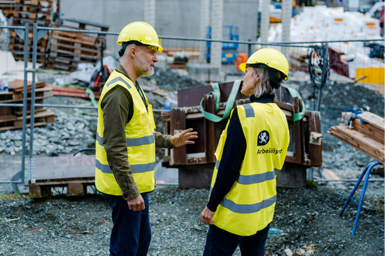 To inspektører med vernehjelm og refleksvest i samtale med hverandre på en byggeplass