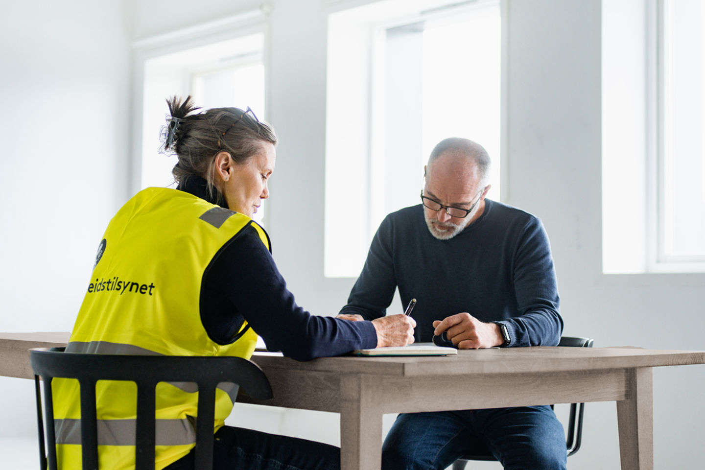 Kvinne og mann i et rom rundt et bord mens hun noterer, han leser