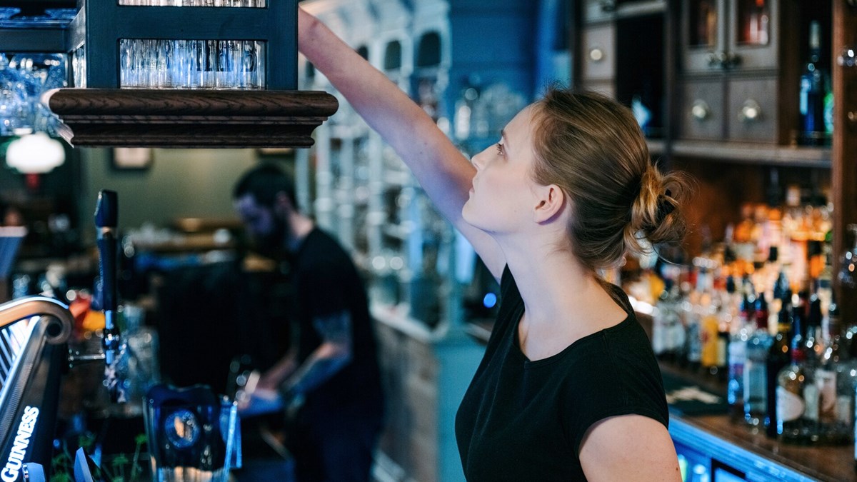 Bildet viser en kvinnelig servitør som strekker seg etter et glass i en bar. 