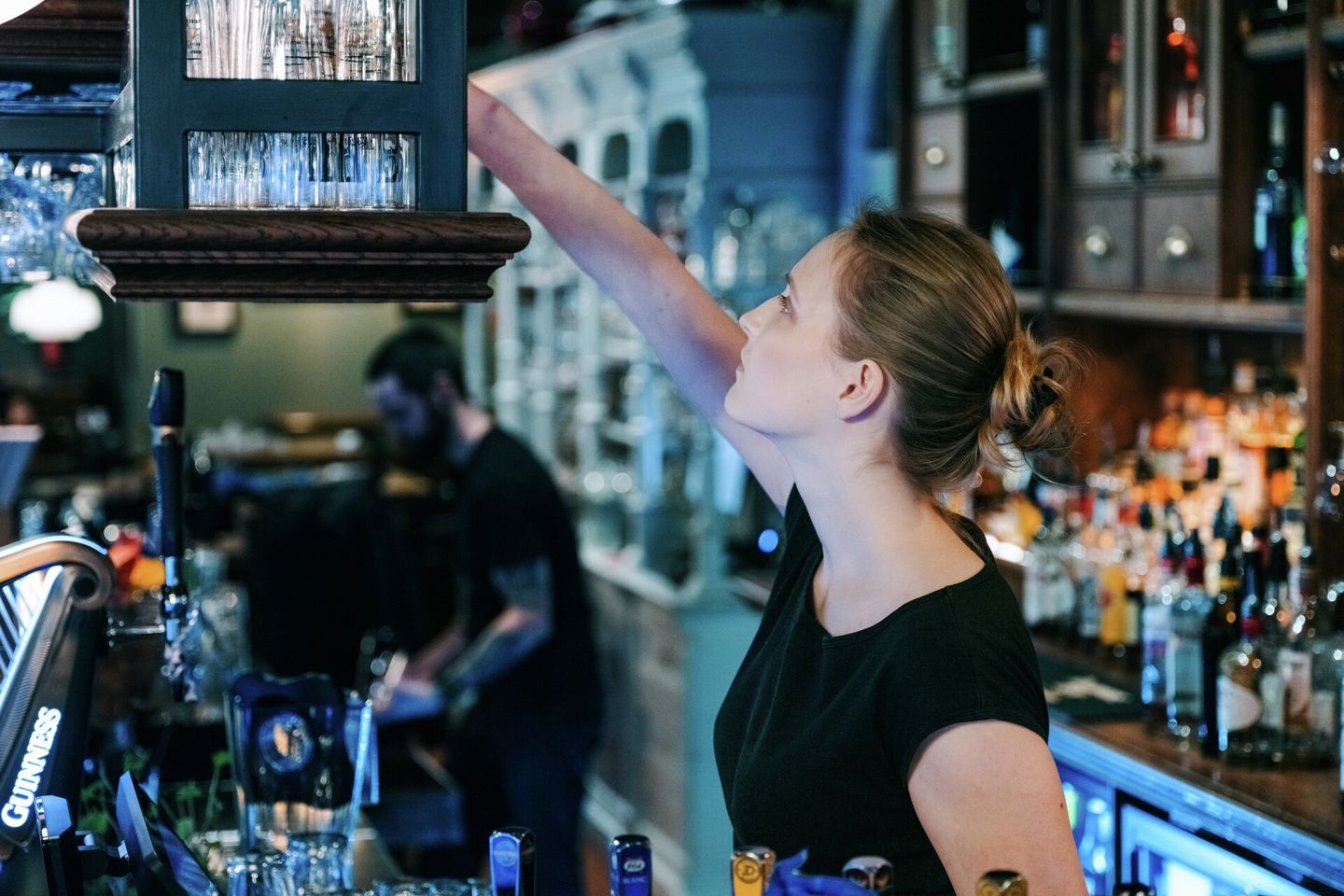 Bildet viser en kvinnelig servitør som strekker seg etter et glass i en bar. 