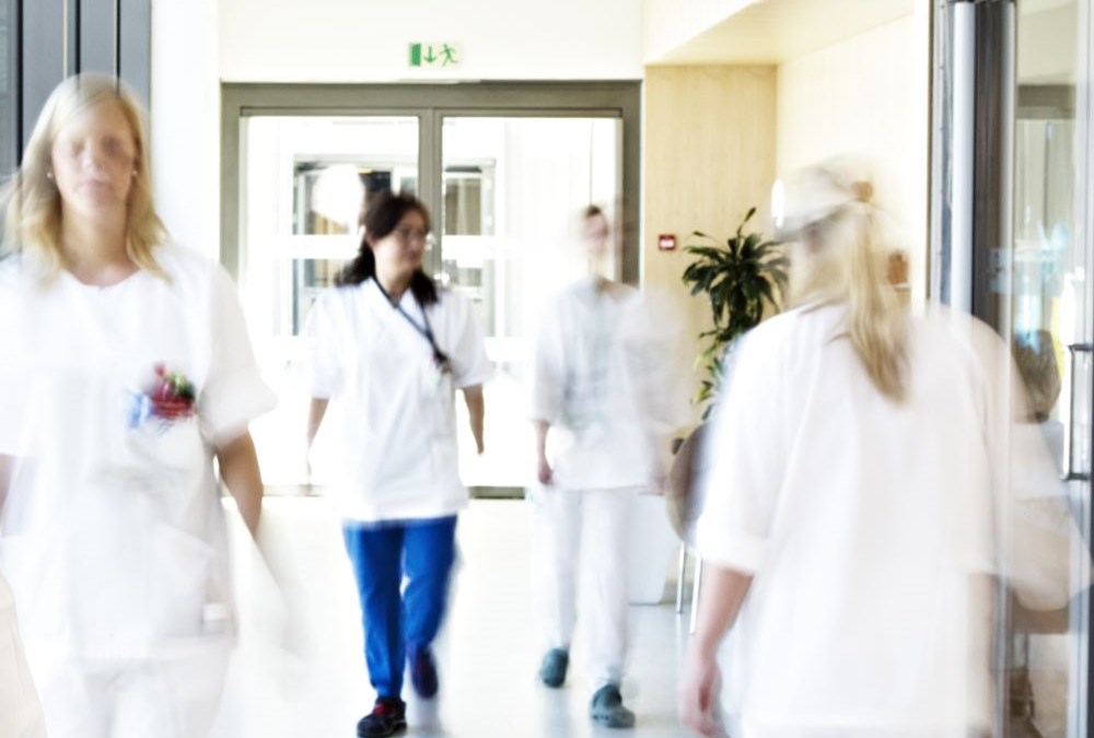 Bildet viser en sykehuskorridor og flere hvitkledde ansatte som går gjennom den. Alle er uskarpe, så man ser ikke ansiktet til de ansatte. 