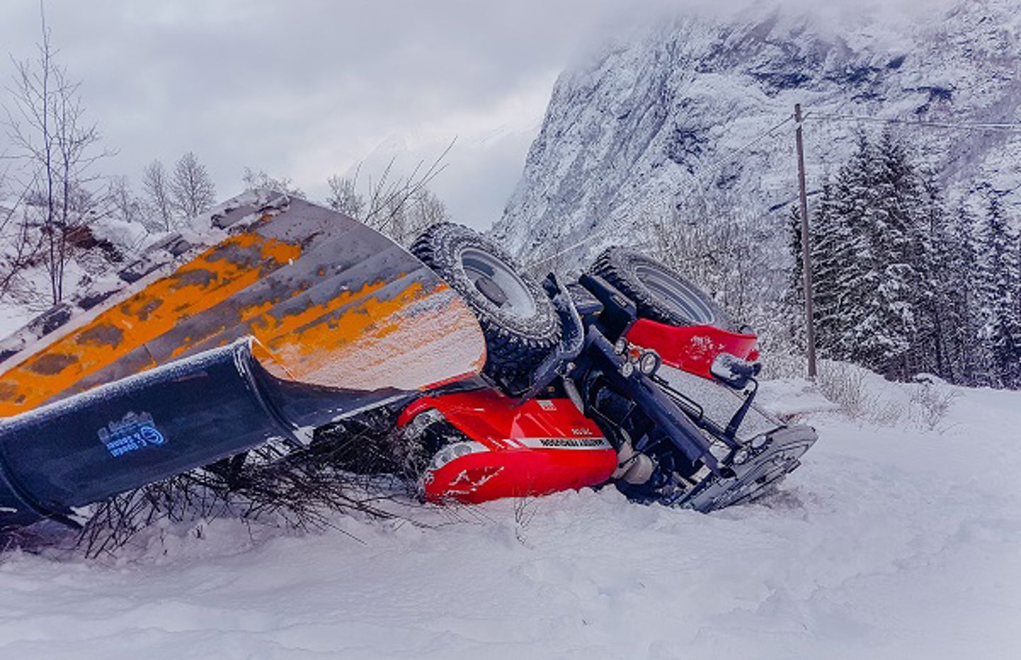 En traktor med plog som har veltet og ligger med hjulene opp. Førerhuset er klemt sammen. Traktoren ligger i snøen. Foto.