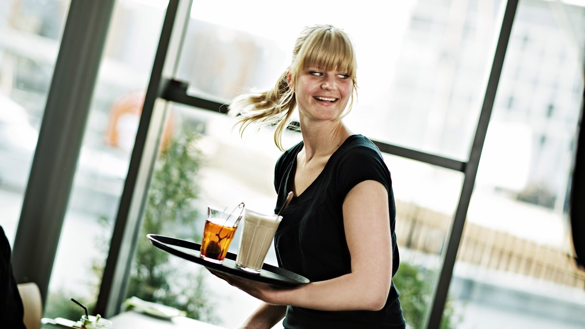 Kvinnelig servitør som ser travel, men smilende ut og holder et brett med en kaffe og en kopp te. Foto.