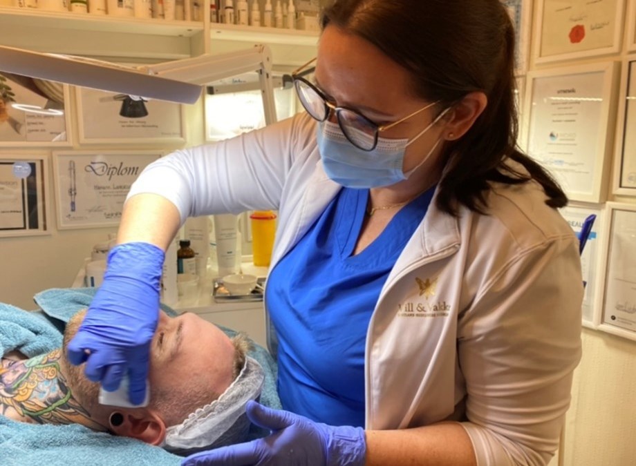 Bildet viser en kvinne med munnbind som utfører ansiktsbehandling på en mann som ligger på en behandlingsbenk. 