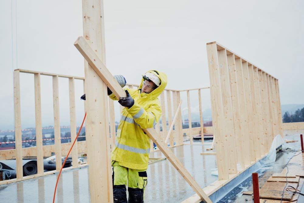 Bildet viser en arbeider som jobber på toppen av et tak. Han holder treverk. 