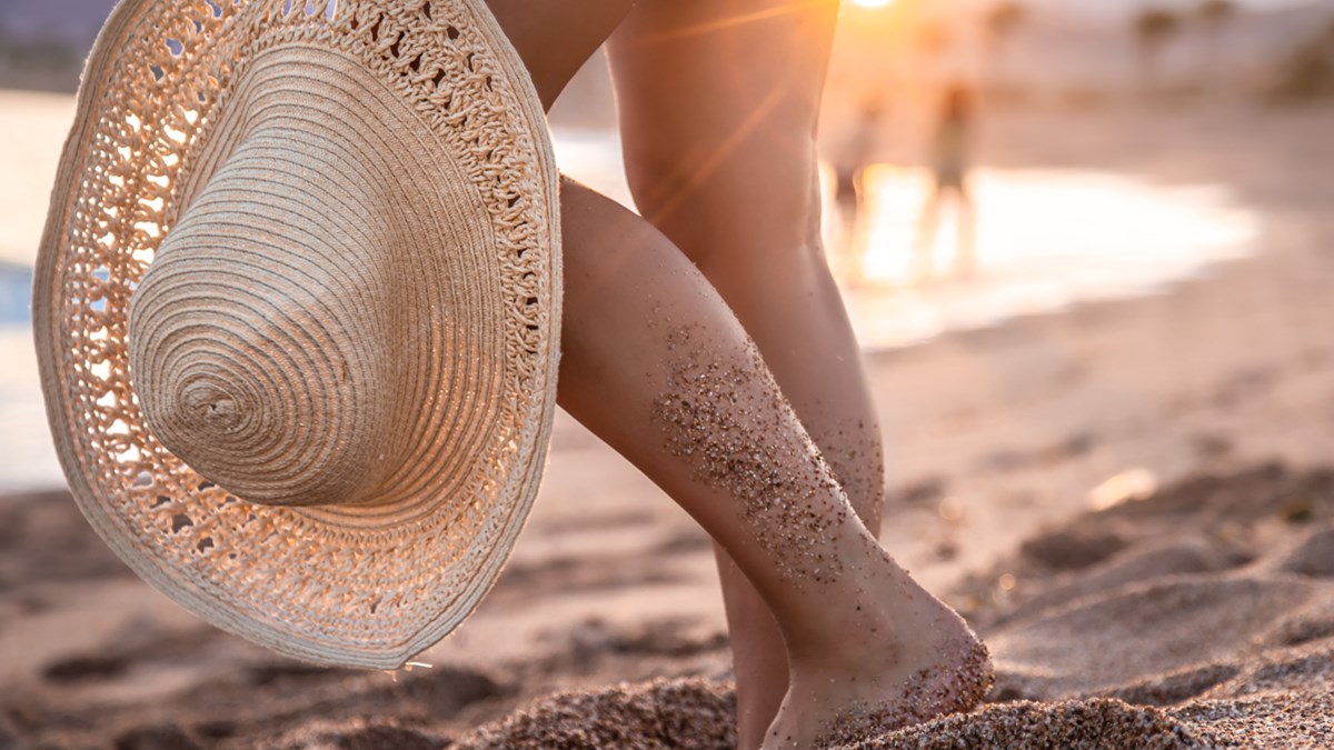 Bildet viser ei hånd som holder ei solhatt og bein og ei strand i bakgrunnen.