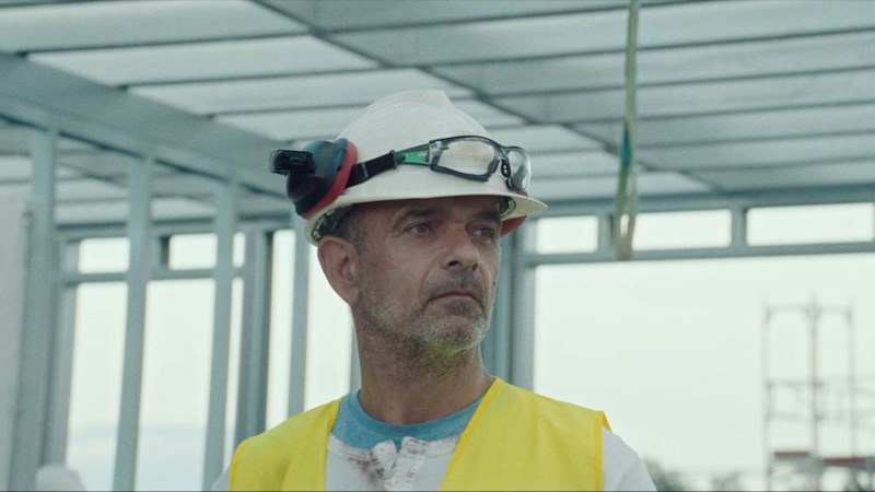Bildet viser en arbeider innen bygg og anlegg med gul vest og hvit hjelm. 