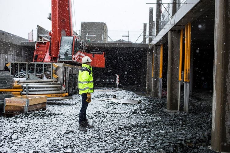 Bildet viser en mann med gul jakke som står på en byggeplass i snøvær.