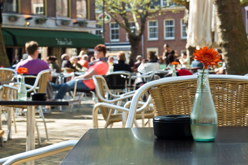 Bildet viser en uteservering på en kafe eller en restaurant. I forgrunnen ser vi en vase med en blomst og et askebeger. 