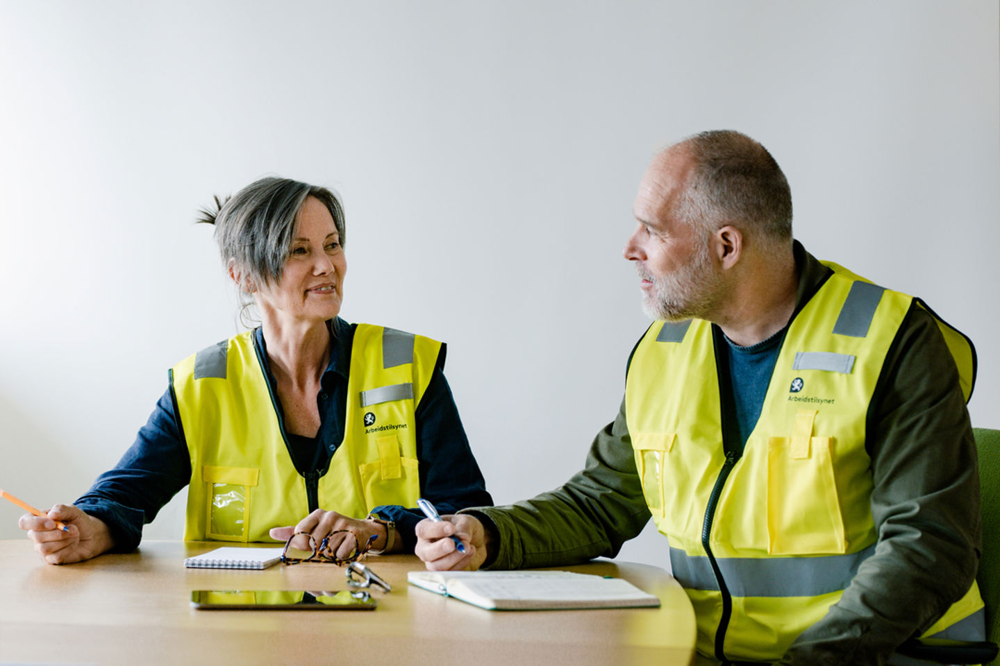 En kvinne og en mann i gule vester har en samtale, papirer foran dem på bordet 