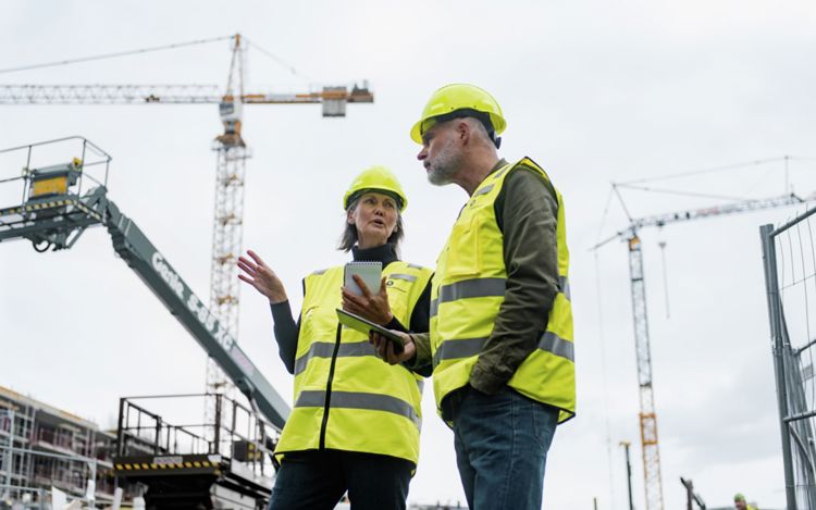 Mann og kvinne med hjelm og refleksvest står og snakker på en byggeplass med to heisekraner i bakgrunnen