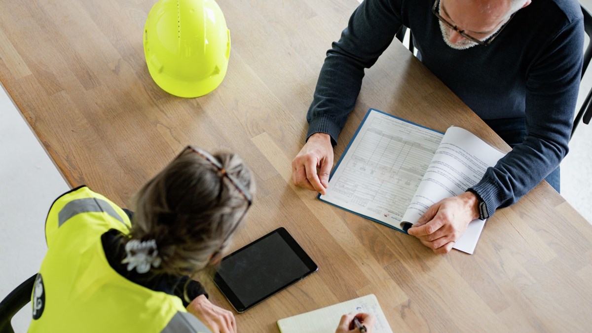 Bildet viser to inspektører i Arbeidstilsynet som sitter rundt et bord og snakker. Begge har på seg gule vester. 